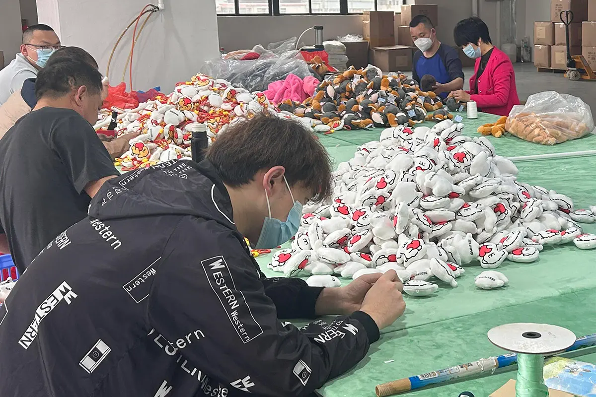 Workers hand sewing and finishing small plush toys on a production table